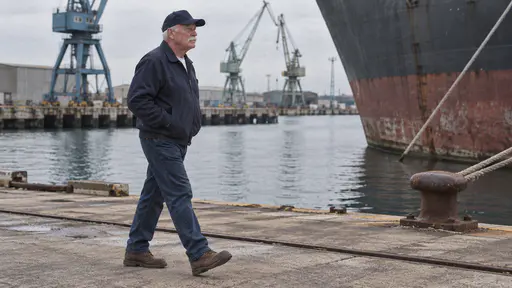 A white veteran in his early sixties in a plain navy cap and jacket walks along a dockyard quay with his hands in his pockets in clear morning light, plain cranes and a moored hull behind him.