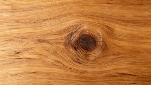 An overhead view of a sanded wooden board in warm honey and amber tones with long flowing grain around a single dark knot under raking light from the left.