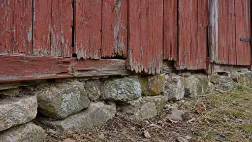 The weathered red boards of a barn wall meet a rough stone footing at ground level with old paint flaking at the joint in soft morning light.