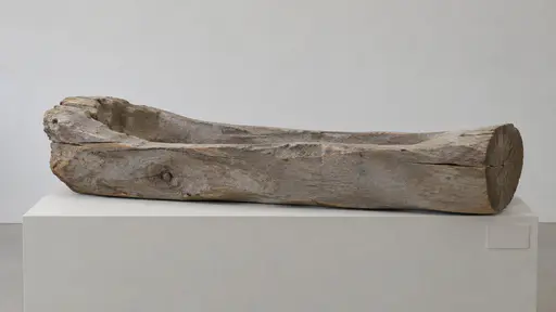 A weathered carved wooden form with greyed surfaces rests on a pale plinth, one end showing clear exposed end grain, against a plain wall.