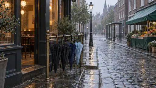 A row of closed black, navy and pale blue umbrellas hooked over a brass railing beside a lit café doorway drip into small puddles on wet cobbles while steady rain falls past a lamp post.