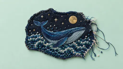 Hand-embroidered textile slice of a whale swimming through French-knotted night waves under a pale gold thread moon, its frayed fabric edge on a pale seafoam field.