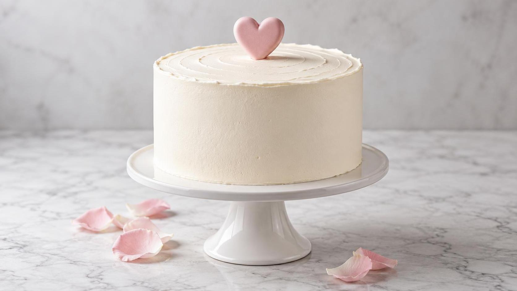 A round white celebration cake with smooth buttercream frosting and a small heart shaped cake topper stands on a cake stand beside a few pale pink petals against a soft bright background.