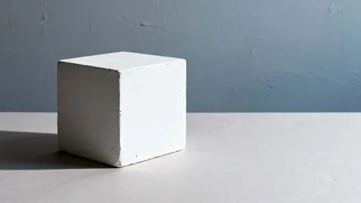 A minimalist white cube sits on a neutral surface within a 16:9 frame, casting a soft, elongated shadow that sculpts the space and emphasizes form and subtle light gradients.