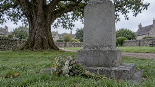 A small bunch of wildflower stems tied with plain twine lies at the foot of a grey stone memorial on a low plinth on a village green with dew on the grass and an old oak behind in cool first light.