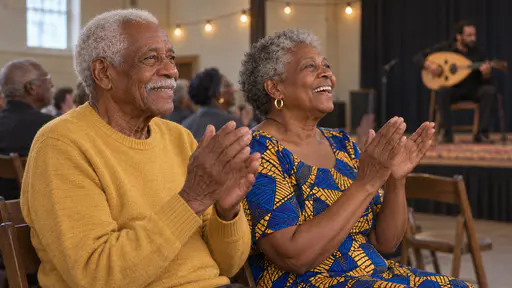 A tall elderly man in a mustard sweater and an older woman with silver curls in a blue and yellow wax-print dress sit in the front row applauding with both hands joined, beaming, with the oud player blurred on the low stage behind.