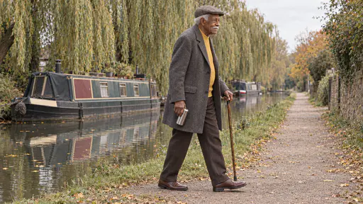 A tall elderly man in a flat cap and grey coat walks along a canal towpath carrying a steel flask and a walking stick beside still water, a moored narrowboat and willows in soft golden light.