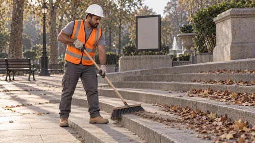 A broad strong man with brown skin and a short black beard in a white hard hat and orange high-visibility vest sweeps the wide stone steps of a park plaza with a broad broom, half the steps swept clean and half still covered with leaves in long evening shadows.